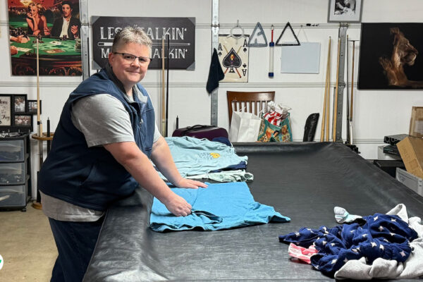 Melia Long folding laundry on a pool table in her garage, surrounded by clothing and household items