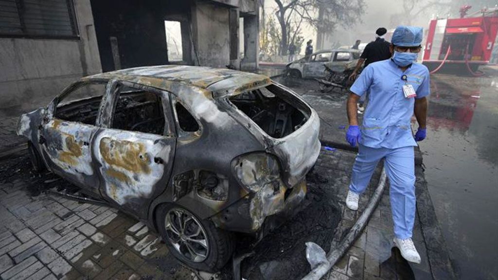 A man in medical scrubs walks beside a burnt out car